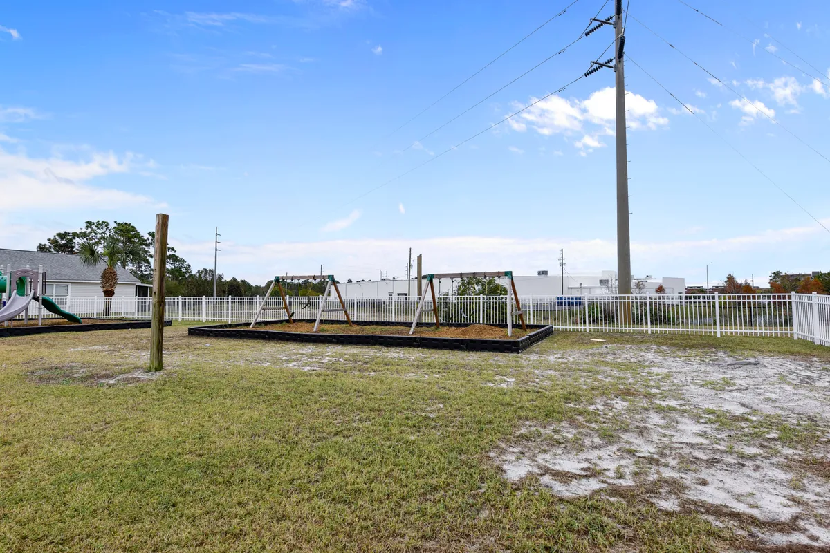 A sunlit, fenced playground with colorful equipment, including swings and slides, surrounded by palm trees and green grass. Families and children are visible, enjoying playtime and picnics in a safe, cheerful environment.