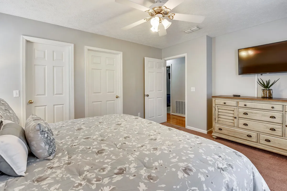 A spacious bedroom featuring a bed with floral bedding, a wooden dresser, a wall-mounted TV, and doors leading to other spaces. The room is filled with soft natural light, creating a comfortable and inviting atmosphere.
