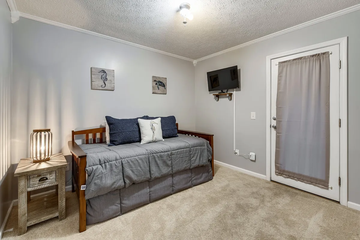A cozy bedroom with a daybed layered in decorative pillows, a nightstand, wall art, and a TV. The room exudes comfort and is ideal for reading, napping, or watching TV—perfect for kids or grandparents.