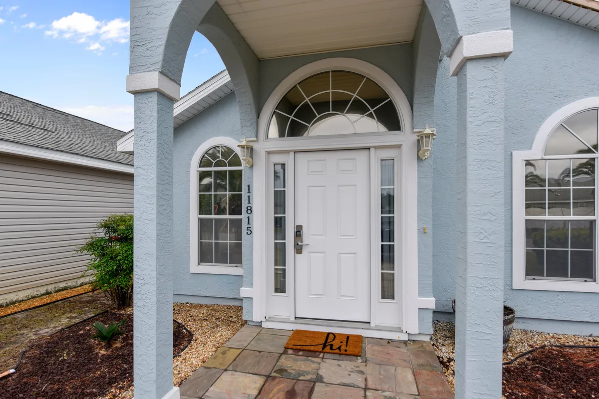 The welcoming front door of the property, framed by landscaped gardens and arched windows. A welcome mat sits at the entrance, suggesting a homely and pet-friendly vibe.