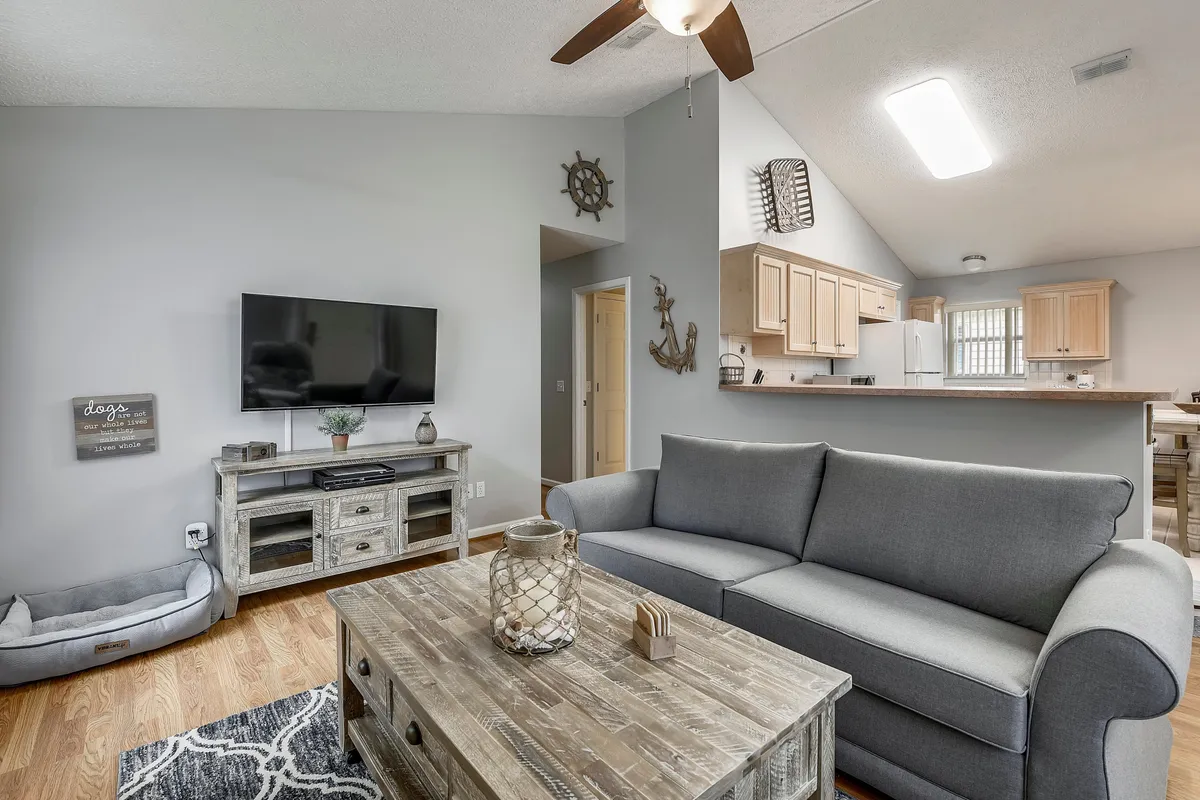 A cozy, open-concept living room featuring a comfortable gray sofa, a rustic wooden coffee table with a decorative lantern, a wall-mounted flat screen TV, and a modern kitchen with bar seating visible in the background. The space is bathed in natural light, and a dog bed can be seen, hinting at pet-friendly amenities.