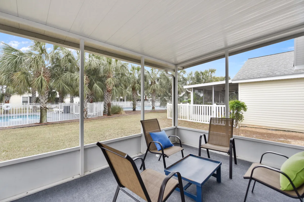 A screened patio seating area furnished with four chairs and a small table, offering a comfortable space to relax outdoors. Beyond the patio, the shared community pool is visible, surrounded by palm trees and adjacent residential homes, all enclosed by a white fence.