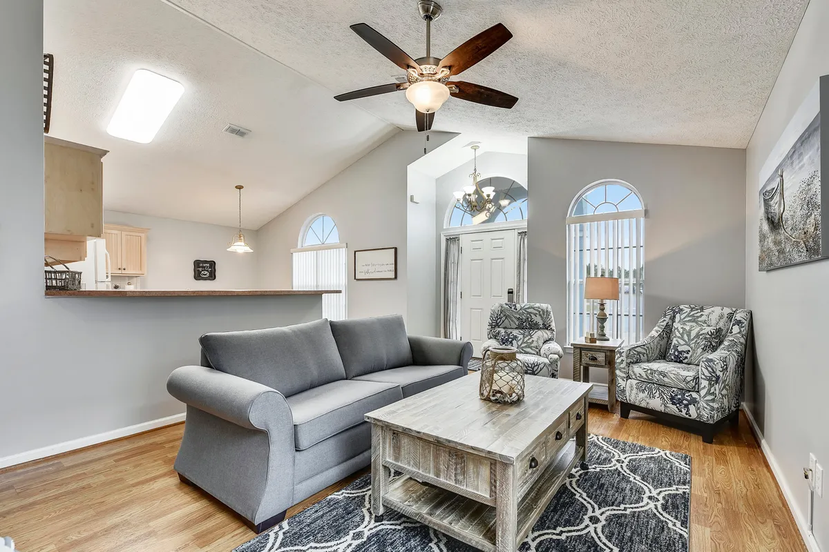 A welcoming living area with a grey upholstered sofa, a wooden coffee table with a distressed finish, two patterned armchairs, and arched windows with vertical blinds providing abundant natural light. The arrangement encourages conversation and relaxation.