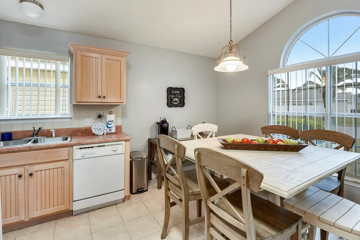 A warm, coastal-inspired dining and kitchen area featuring a wooden dining table with a fresh fruit centerpiece, a large arched window with bars offering a glimpse outside, a compact kitchen with a dishwasher and sink, and a corner coffee station for morning rituals.