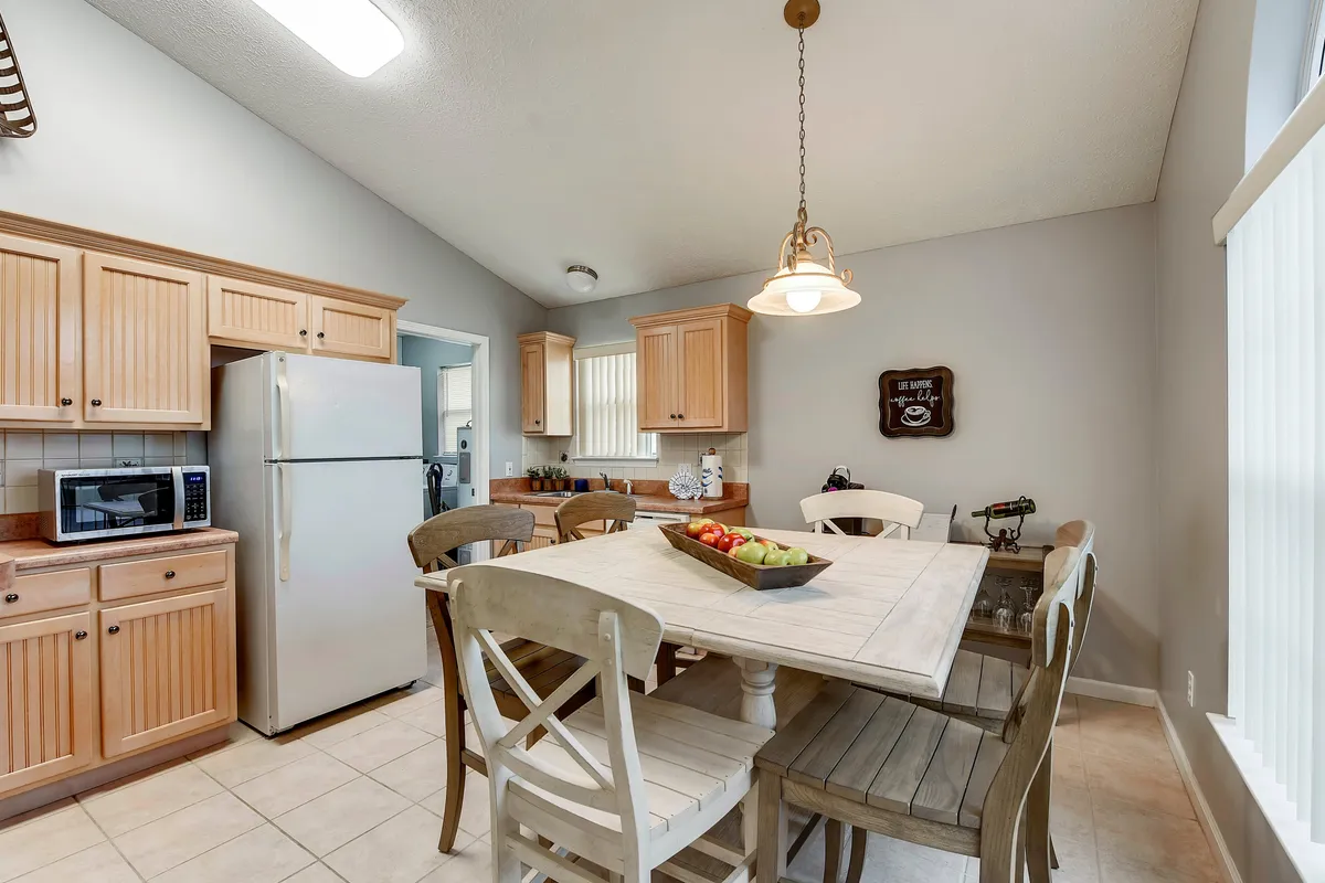 A cozy wooden dining table with mixed chairs sits beside a kitchen with wooden cabinets and a white refrigerator. Soft lighting and a welcoming wall sign evoke warmth and encourage communal meals or coffee chats.