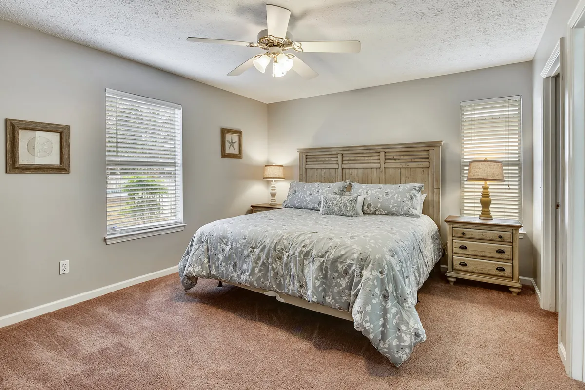 A serene master bedroom featuring a king-size bed with floral bedding, a wooden headboard, matching nightstands, and soft table lamps. The room is softly lit and includes a ceiling fan with lights, exuding comfort and tranquility.