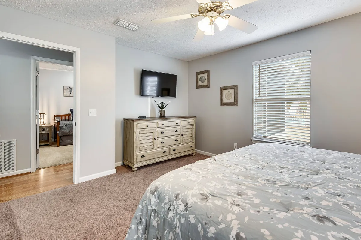 A bright and inviting bedroom with a floral patterned bedspread, wall-mounted flat screen TV, wooden dresser with a plant, and a window letting in natural light through blinds. The setup echoes hotel-style comfort and relaxation.