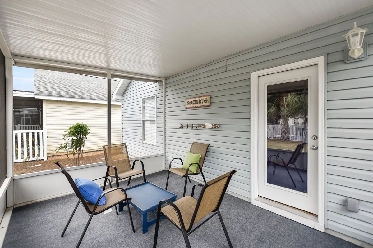 A cozy patio area featuring four patio chairs with colorful cushions, a small blue table, and a decorative 'seaside' wall sign. The space is adjacent to a glass door, evoking a relaxed, beachside retreat ideal for morning coffee or evening relaxation.