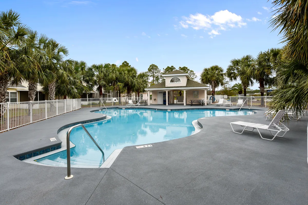 A large, inviting community swimming pool with clear blue water, lined with lounge chairs and shaded by tall palm trees. There is a covered pavilion with picnic tables, and the area is enclosed by a white safety fence, all set in a tranquil, tropical environment.