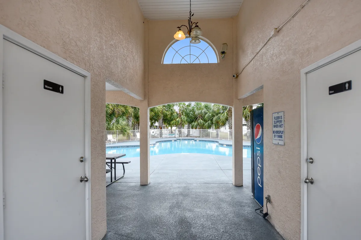 A sunlit community swimming pool surrounded by palm trees, with a picnic table in the foreground and restroom facilities nearby. The area is clean and inviting, ideal for poolside lounging or social gatherings. A Pepsi vending machine adds a touch of convenience for guests enjoying a day by the pool.