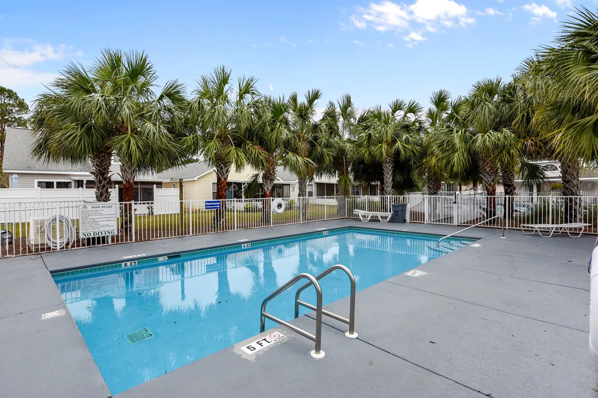 A relaxing community swimming pool is showcased here, encircled by palm trees and a white safety fence. Poolside lounge chairs offer a spot to unwind, with nearby single-story houses visible in the background. The setting is tranquil, family-friendly, and ideal for keeping an eye on kids while relaxing.