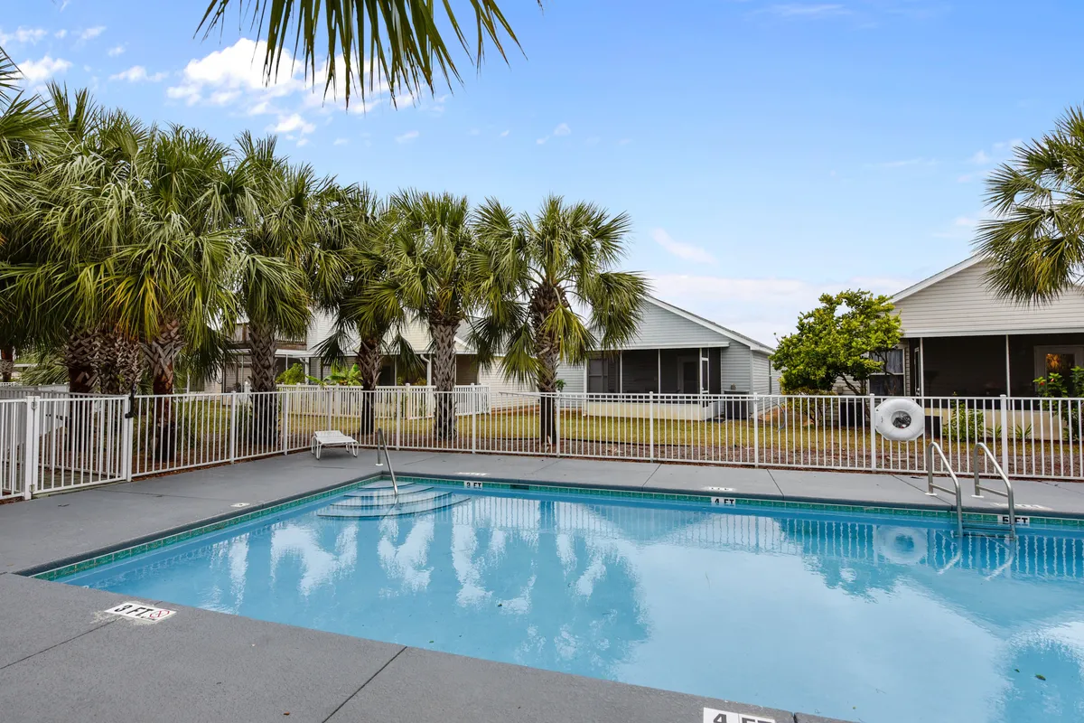 A sparkling clear swimming pool is surrounded by a white safety fence and lush palm trees. Nearby, single-story houses with screened porches overlook the pool. The space feels secluded yet open, ideal for families and groups to gather while children play safely in the water.