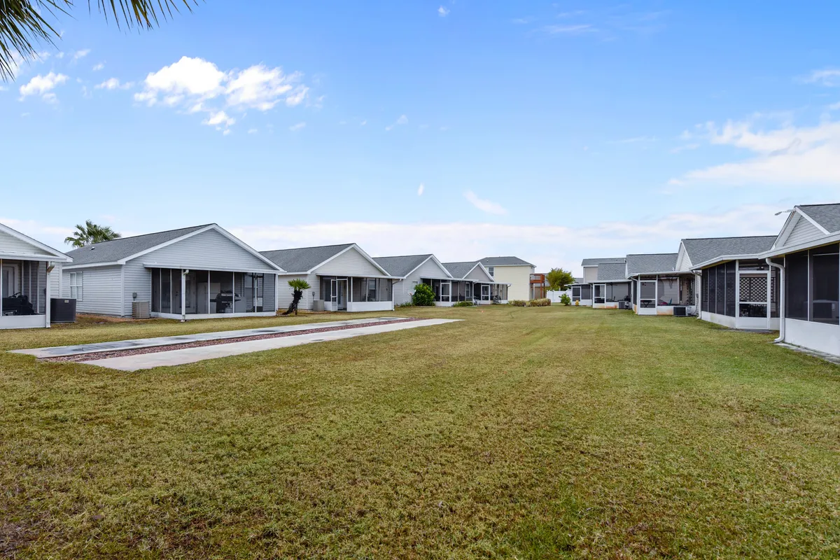 A row of cozy, single-story houses with screened porches lines a large central grassy lawn. Two bocce ball courts or similar lawn games are visible, creating a sense of community and outdoor leisure. The open, green space is ideal for relaxed walks, pet playtime, or casual gatherings with neighbors.