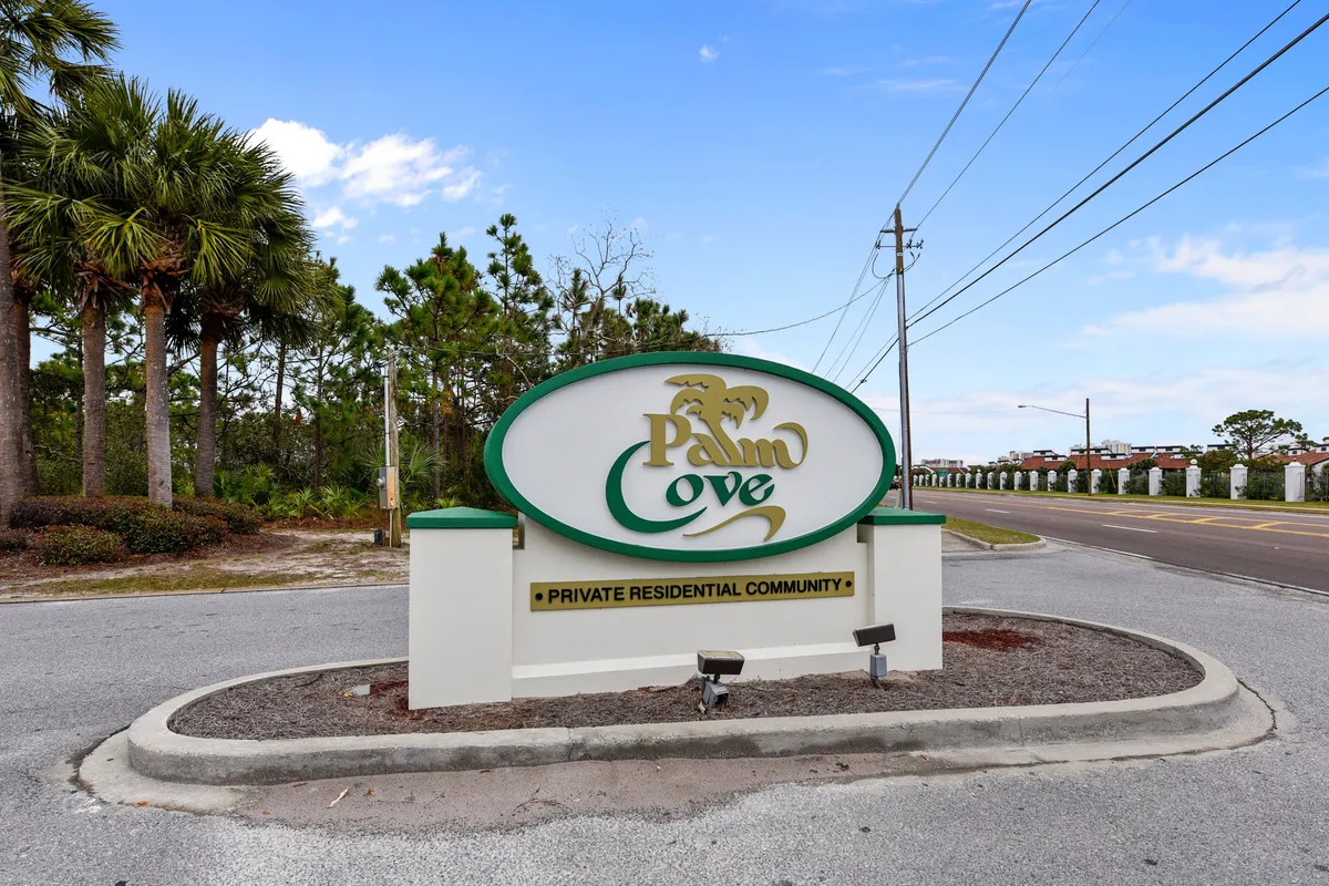 A welcoming signpost marks the entrance to the Palm Cove private residential community, with a paved road inviting visitors in. Tall palm trees and lush greenery surround the entrance, and a clear blue sky enhances the tranquil, inviting atmosphere. The road hints at walkable paths and a safe, pleasant neighborhood for strolls with pets.