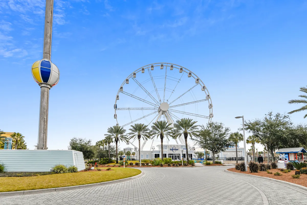 A large Ferris wheel stands over a landscaped path lined with palm trees, near the SkyWheel entrance building. The area is family-friendly and bustling, with space for walking and sightseeing, and hints at lively seasonal events and amusement rides.