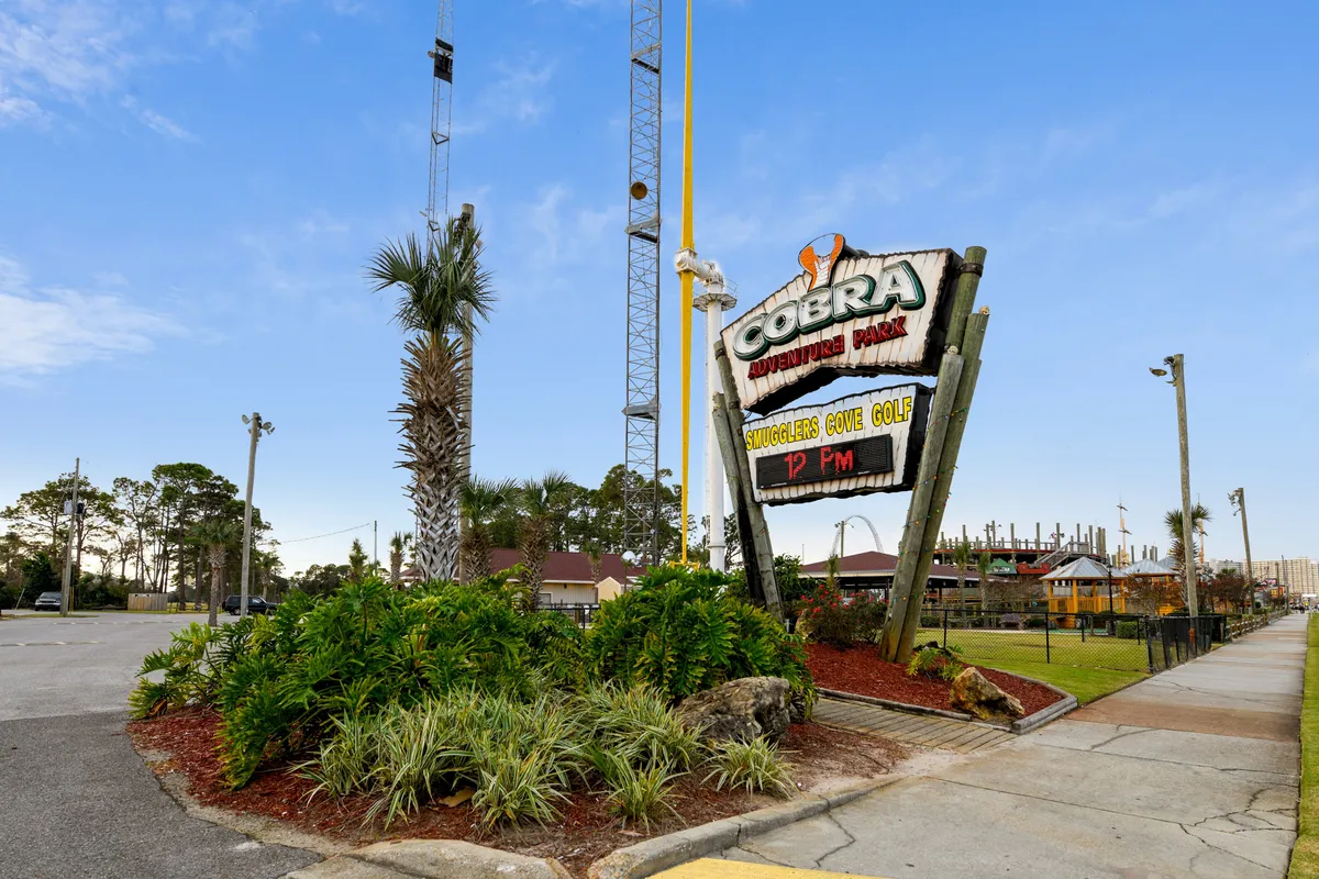 The image features the entrance sign for Cobra Adventure Park, with tall amusement rides visible in the background and lush greenery with palm trees framing the scene. A sidewalk leads visitors past the park entrance, promising a day of excitement and friendly competition with mini-golf and thrill rides.