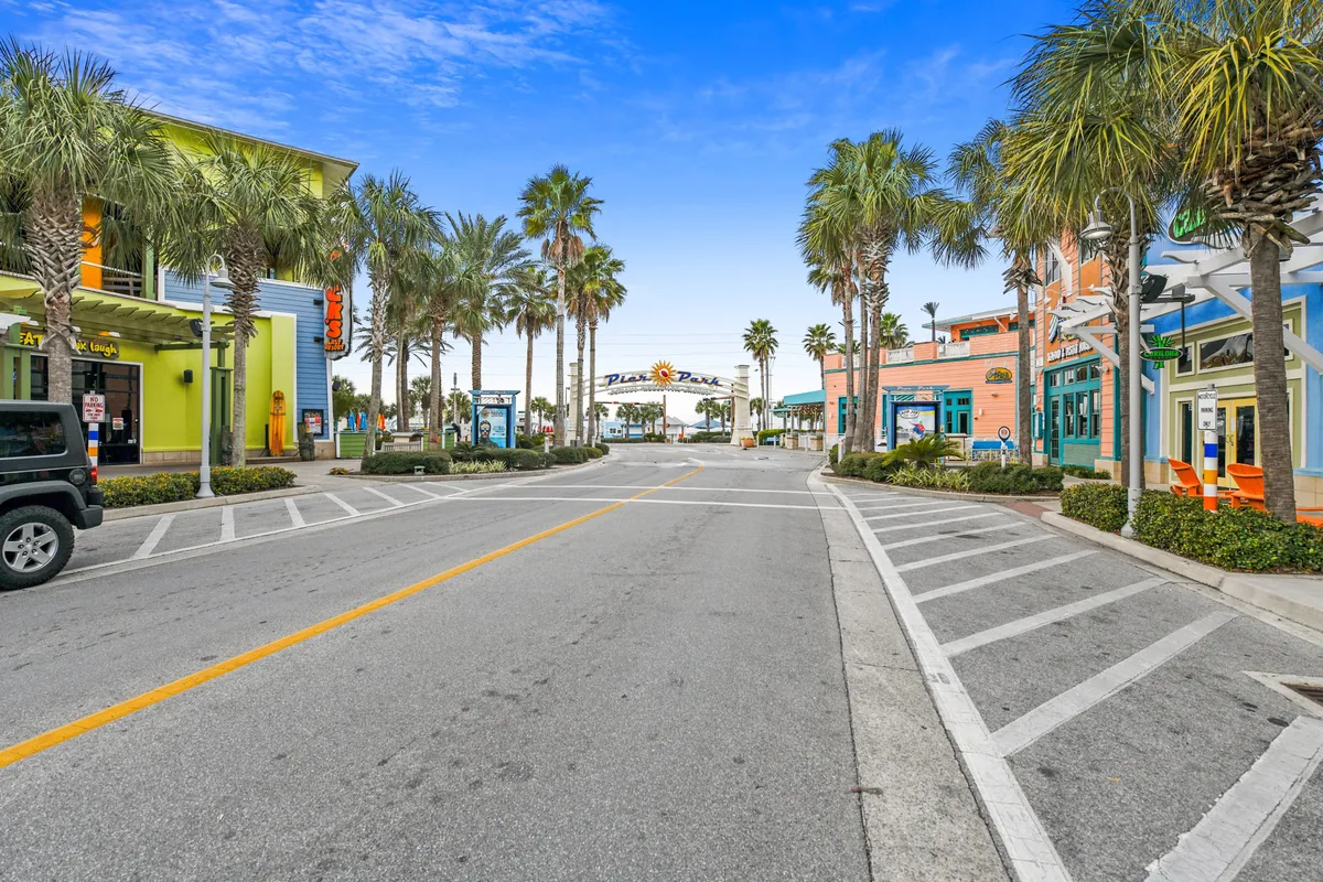 A lively street scene at Pier Park, featuring palm-lined sidewalks, vibrant storefronts, and the iconic Pier Park sign in the distance. The area is bustling with opportunities for shopping, dining, and leisurely strolls, evoking a cheerful beach town ambiance.