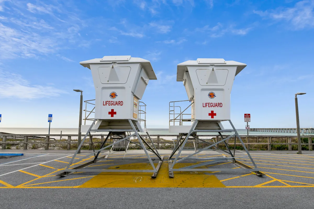 This image features two lifeguard towers in the foreground, with the vast sandy beach and the ocean stretching out beyond a wooden fence. The iconic pier extends into the ocean on the right side, all beneath a bright blue sky with scattered clouds. The area is clearly set up for safe, fun beach days with family.