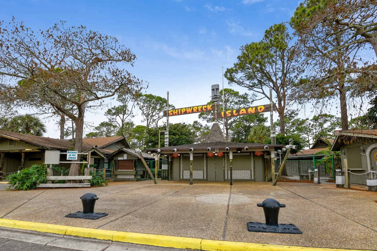 The image showcases the entrance signage to Shipwreck Island, a popular water park in Panama City Beach. Rustic wooden structures and cabins frame the paved entrance area, surrounded by tall trees, evoking a sense of adventure and outdoor fun. The open space in front of the entrance hints at the anticipation and excitement of family outings and group activities.